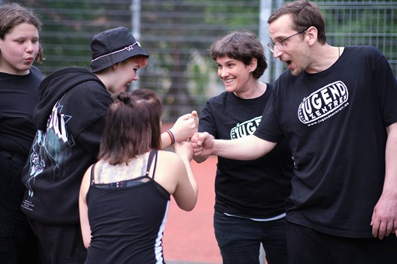 © Mili Badic Jugendliche sitzen vor einem Basketball-Platz, während Jugendarbeiter:innen mit ihnen sprechen