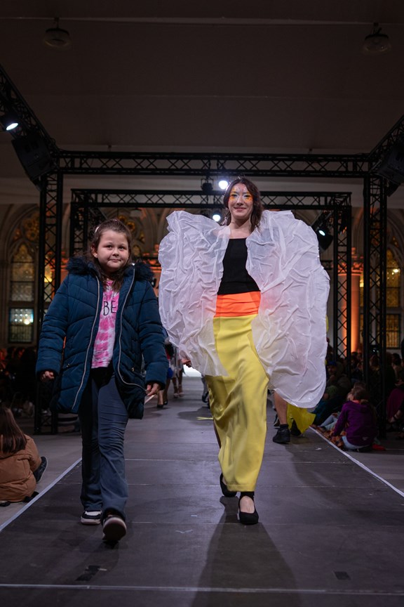© leggou.vision  Kind mit blauer Jacke und blauer Hose neben Model auf dem Laufsteg mit gelber Hose, weißen Schuhen, rotem Gürtel, schwarzem Top und seidigen, weißen Ärmeln