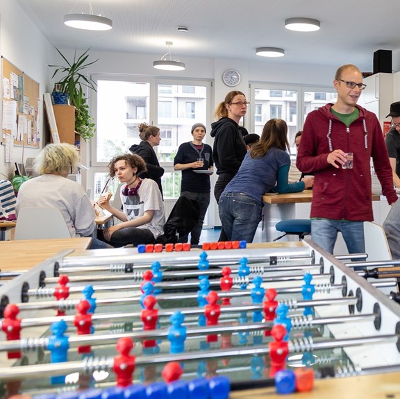 © Verein Wiener Jugendzentren Hinter einem Wuzzlertisch stehen vielen Menschen und unterhalten sich