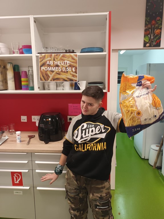 Eine Jugendliche (Michelle) steht in einer Küche und hält einen Sack mit gefrorenen Pommes in die Luft. 