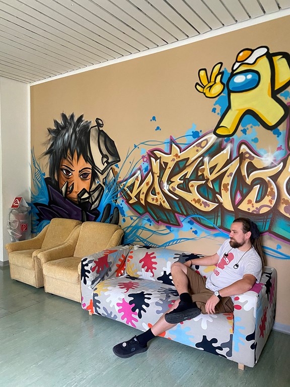 Ein Jugendarbeiter sitz auf einer Couch vor einer Wand, auf der viele Graffitis zu sehen sind. 