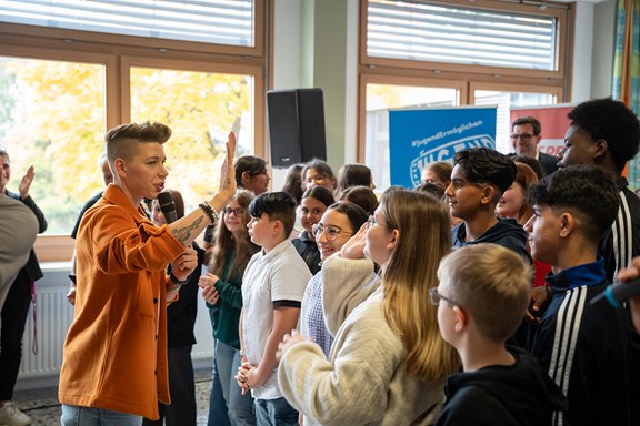Eine Person im orangen Hemd macht ein HighFive-Zeichen zu einer Gruppe von Schüler:innen. 