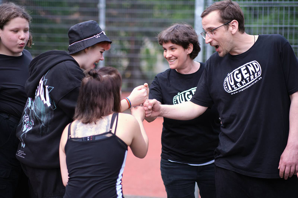 Jugendliche sitzen vor einem Basketball-Platz, während Jugendarbeiter:innen mit ihnen sprechen