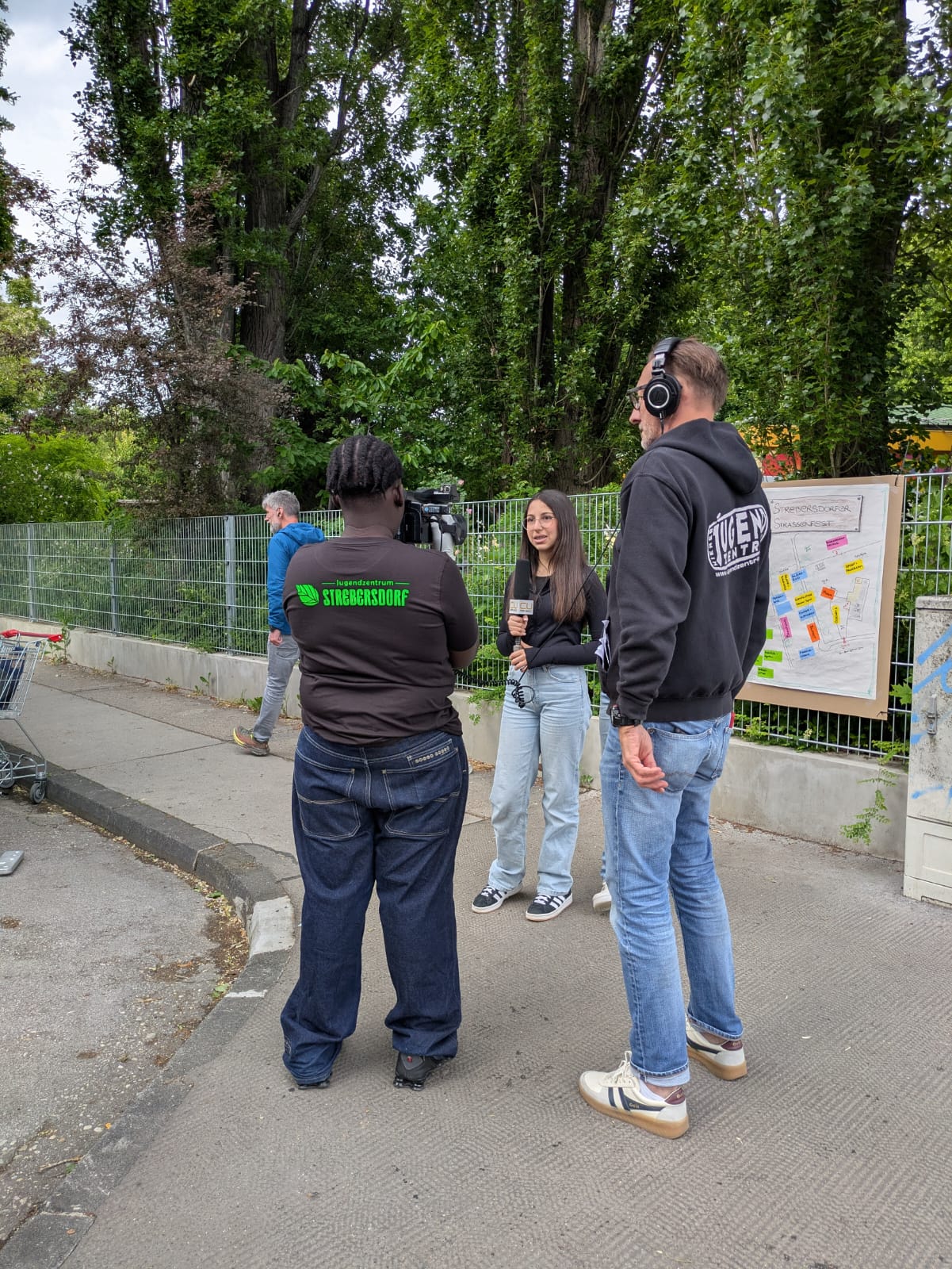 Jugendliche Kayra mit Mikrofon vor der Kamera auf der Straße; hinter der Kamera zwei Kameramänner