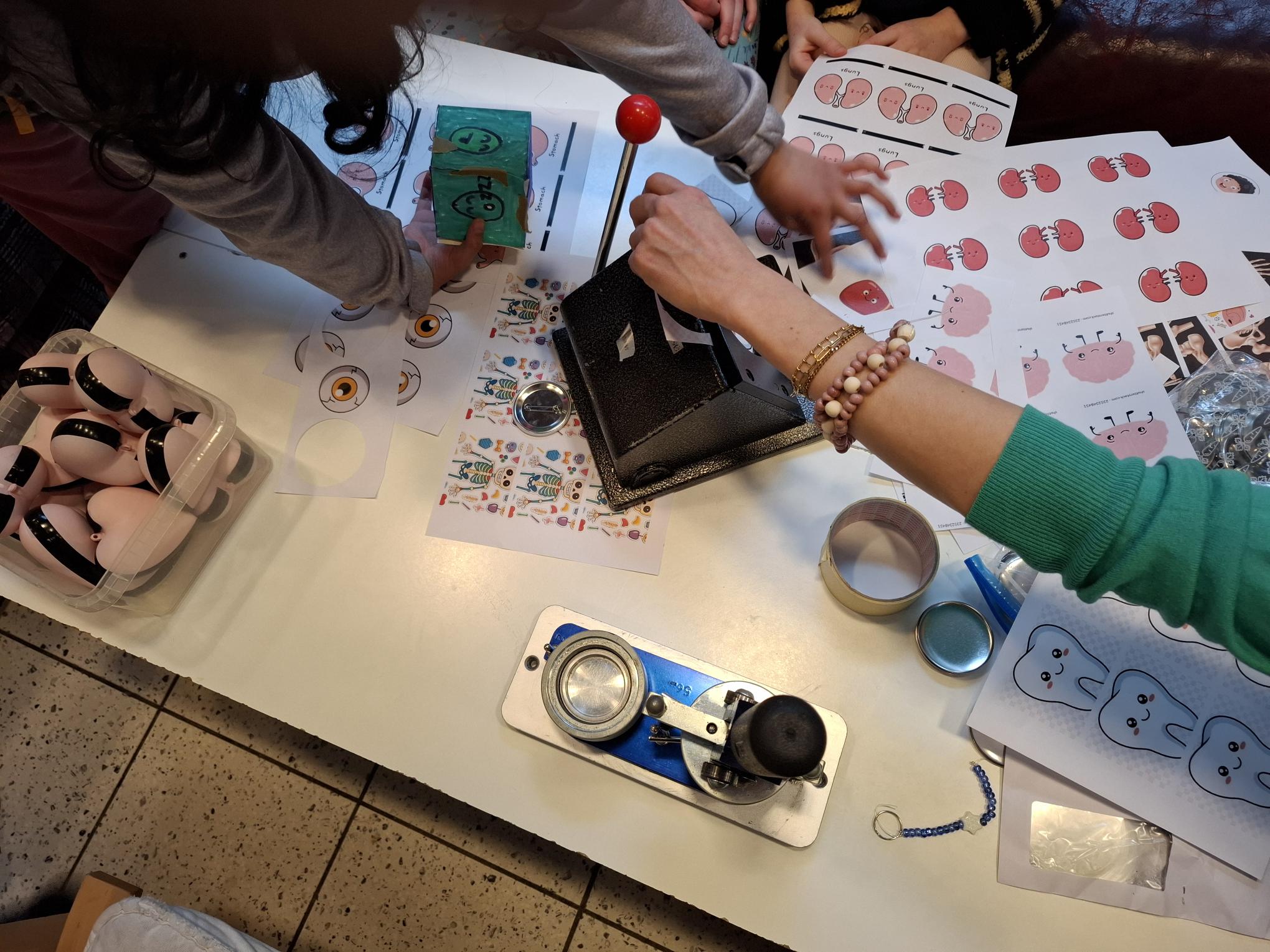 copyright: Auf einem Tisch steht eine Buttonmaschine und Vorlagen auf Papier, auf denen Cliparts von Organen zu sehen sind.