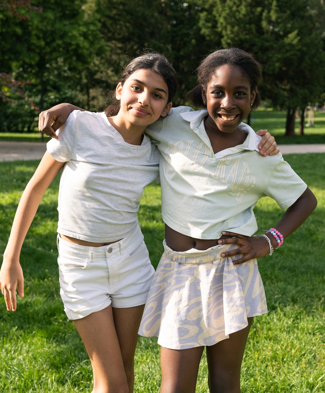 Zwei Mädchen* umarmen sich und lächeln in die Kamera, auf einer grünen Wiese