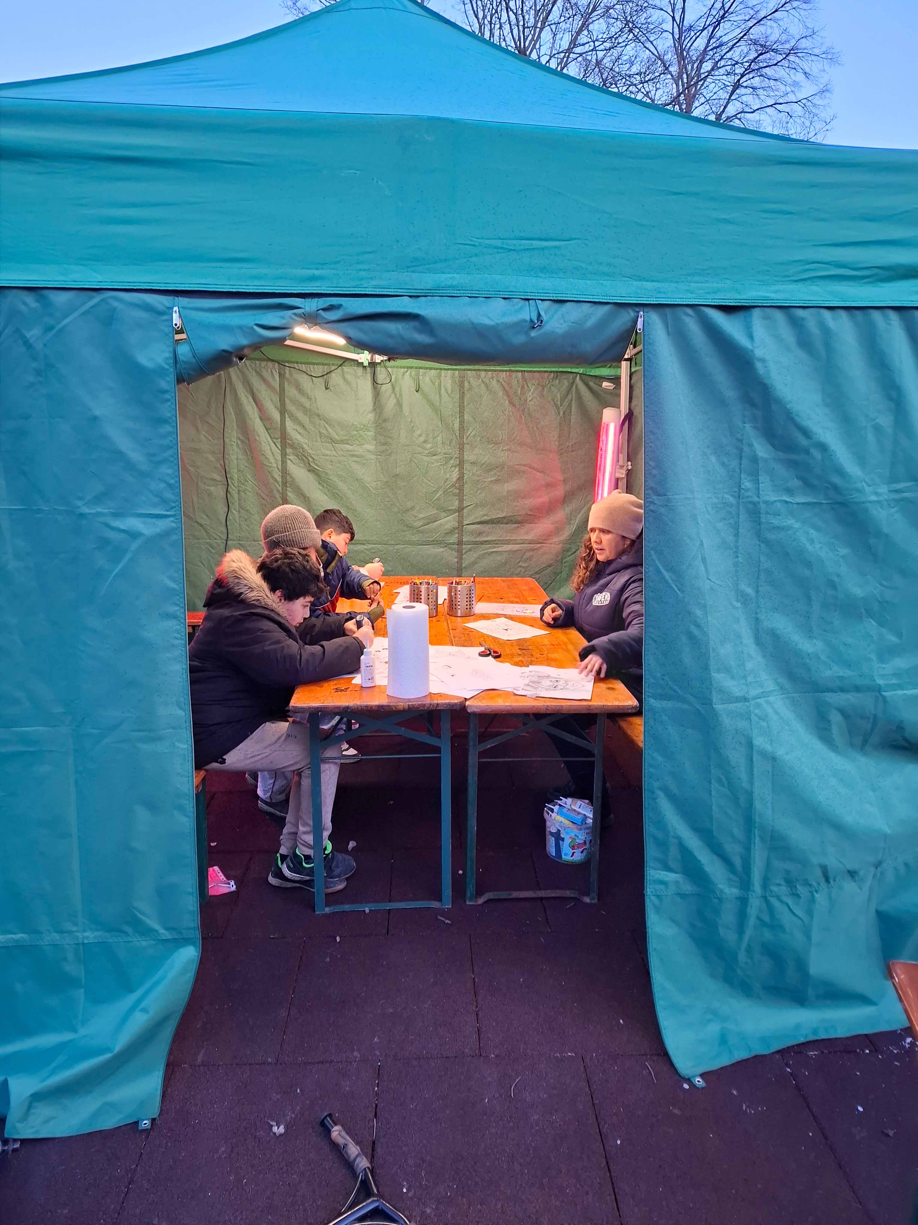 Drei Kinder sitzen gemeinsam mit einer Jugendarbeiterin im Zelt des Jugendzentrums Hirschstetten und basteln.