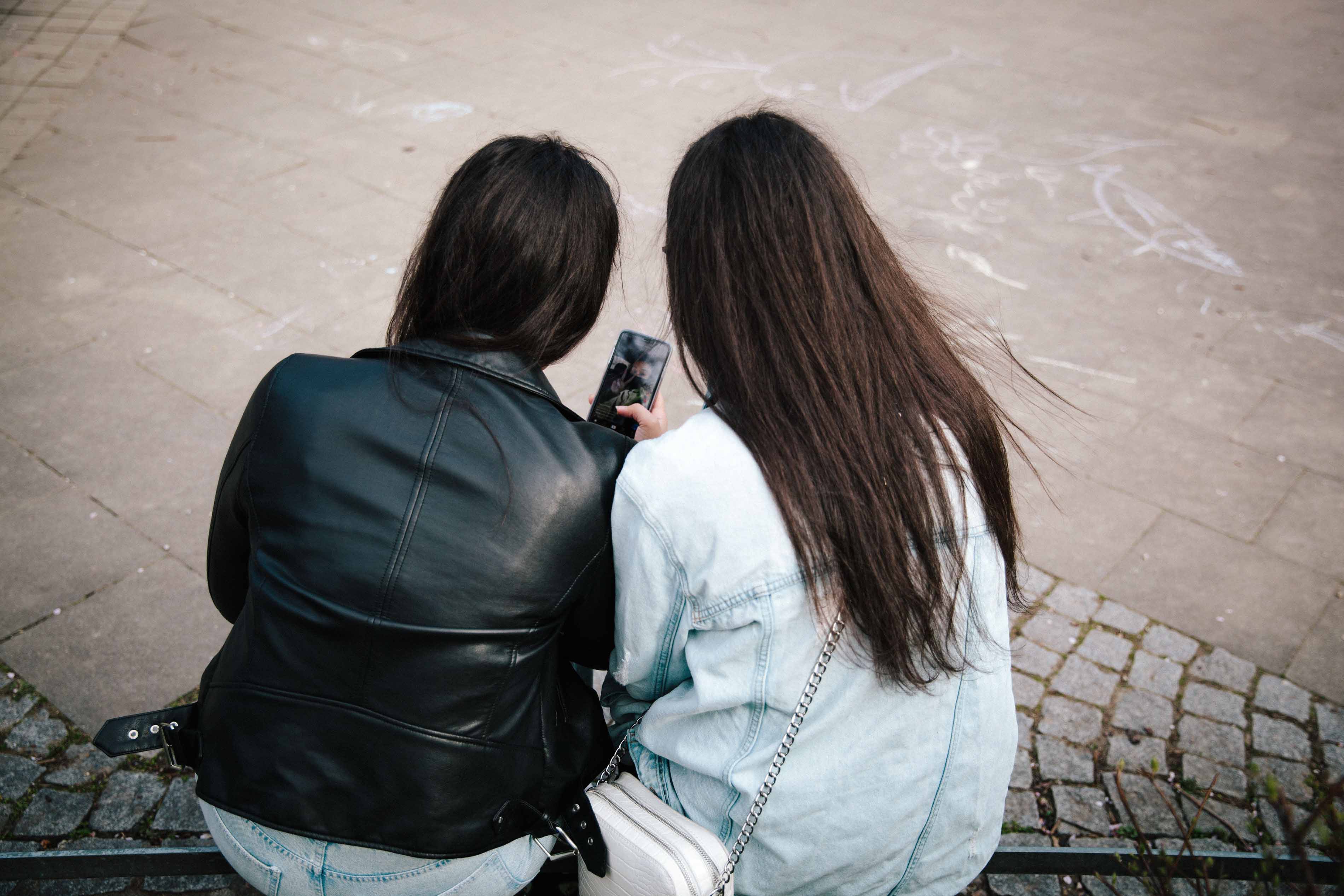 Zwei Mädchen sitzen im Park und schauen gemeinsam aufs Handy.