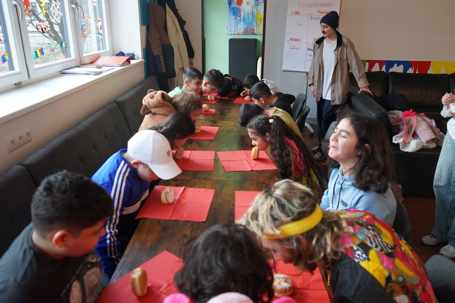 Kinder sitzen um eine Tafel und essen einen Krapfen ohne Hände