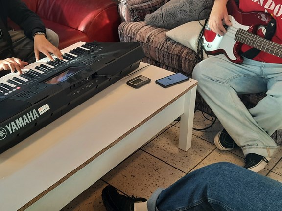 Jemand spielt ein Keyboard, das auf einem niedrigen Tisch liegt. Daneben sitzt eine weitere Person, die einen roten Bass spielt.