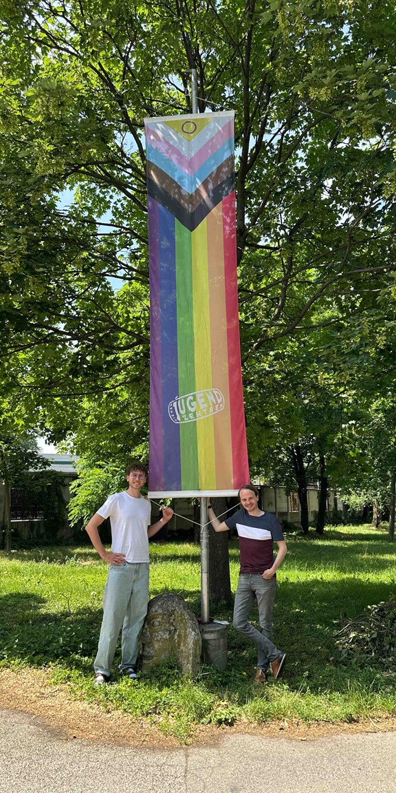 Zwei Mitarbeiter des Vereins hissen Progress-Pride-Flagge beim Eingang zur zentralen Geschäftsstelle des Vereins Wiener Jugendzentren.