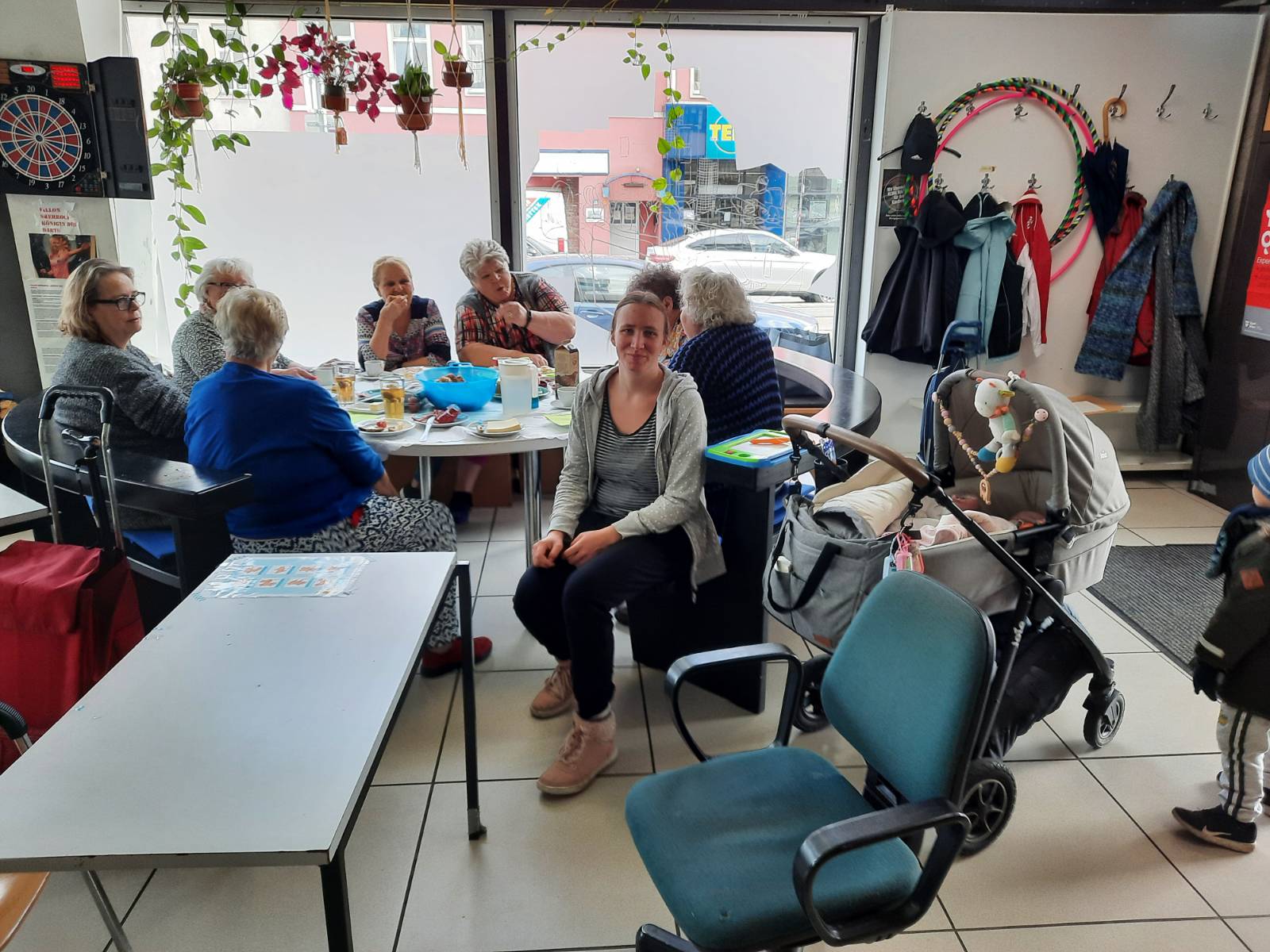 Frauen* sitzen um Tisch mit Jausen und Snacks, Kinderwagen zu sehen und Frau lächelt in die Kamera