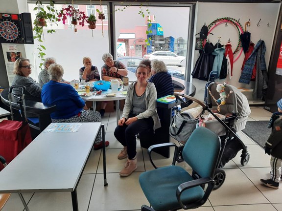 Frauen* sitzen um Tisch mit Jausen und Snacks, Kinderwagen zu sehen und Frau lächelt in die Kamera